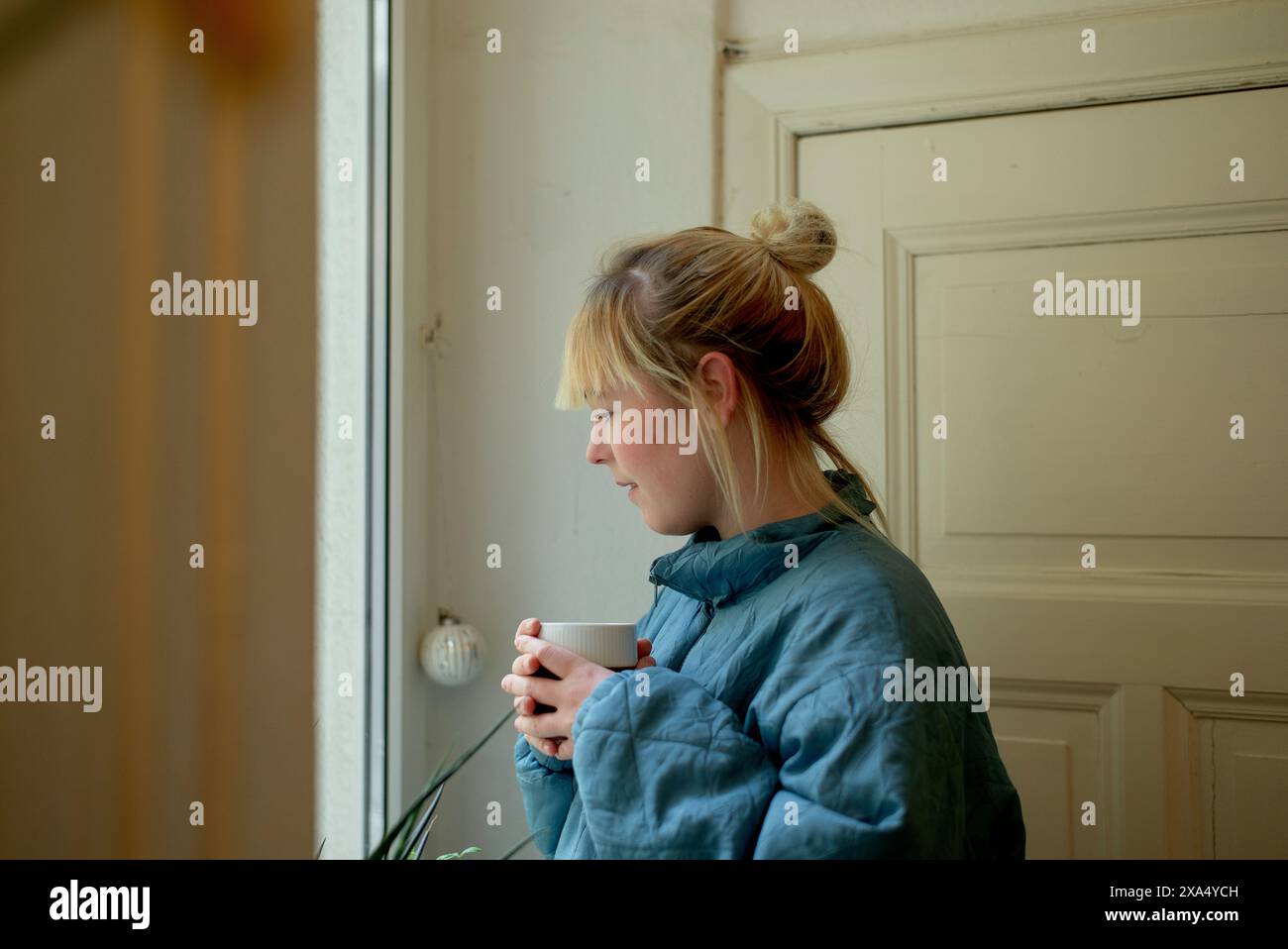 Woman in a blue quilted coat holding a white mug and looking thoughtfully out a window. Stock Photo