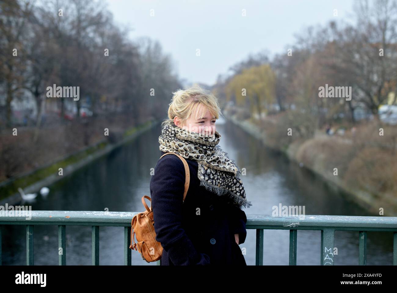 Woman with a scarf and backpack standing on a bridge over a canal on a cloudy day. Stock Photo