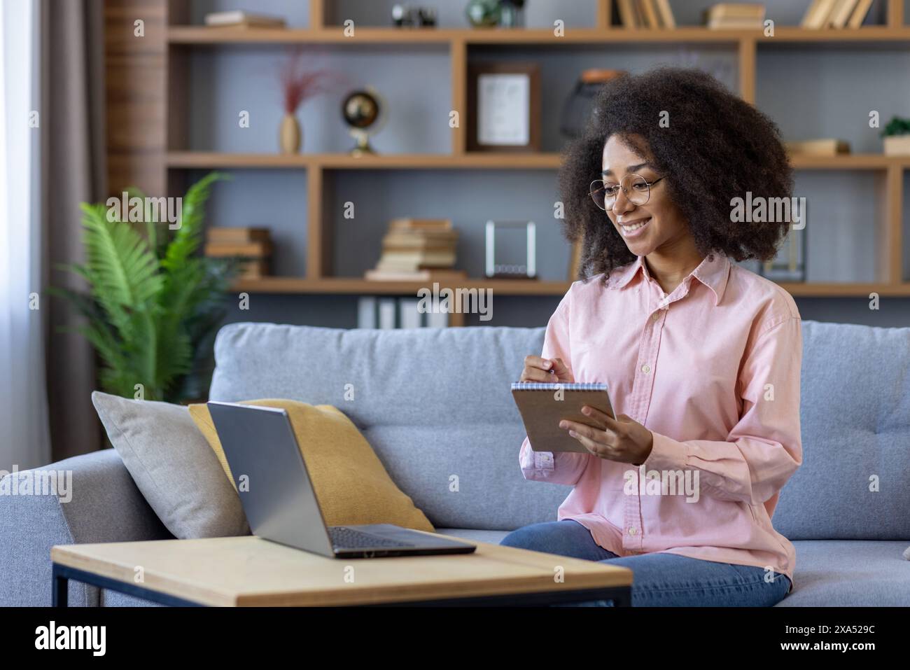 Young student joyfully learning from home, taking notes while engaged in an online video course on laptop. Comfortable home study environment. Stock Photo