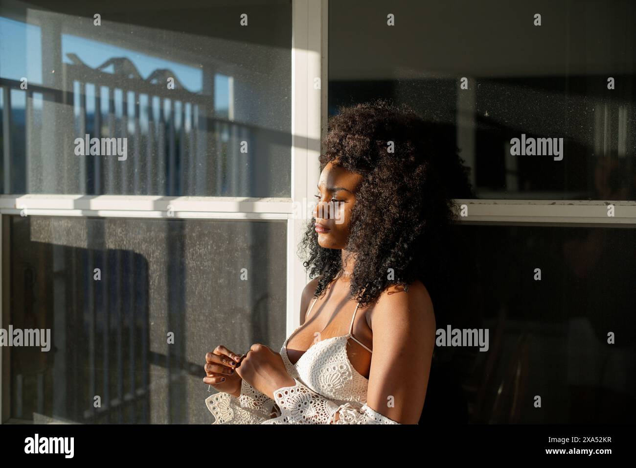 Serene young woman with curly hair enjoys the sunlight by a window. Stock Photo