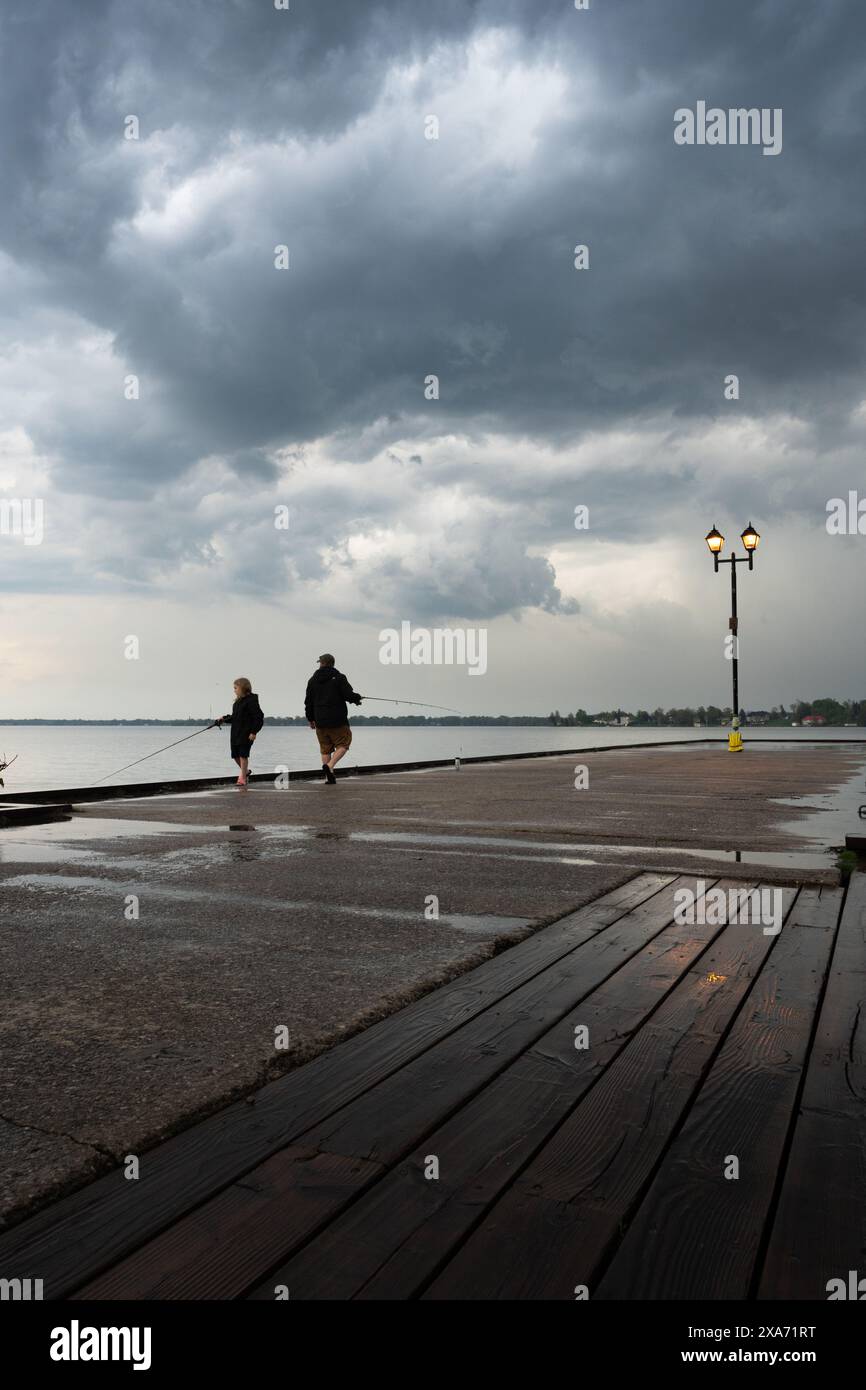 A Father and son fishing off a pier during stormy weather with dramatic clouds over the lake Stock Photo