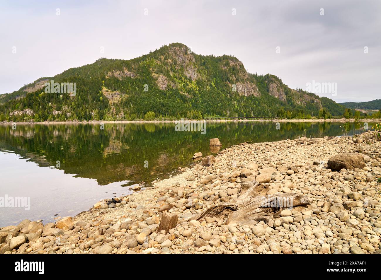 View from a rocky gravel beach of the steep sides of a logged mountain and it's reflection in the calm lake. Stock Photo