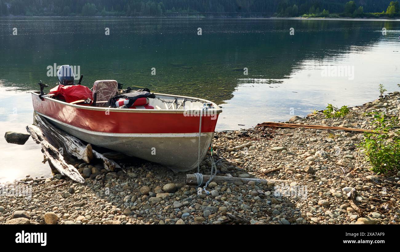 A small red and silver aluminum boat on a gravel beach at the edge of a lake with forest reflections in the lake. Stock Photo