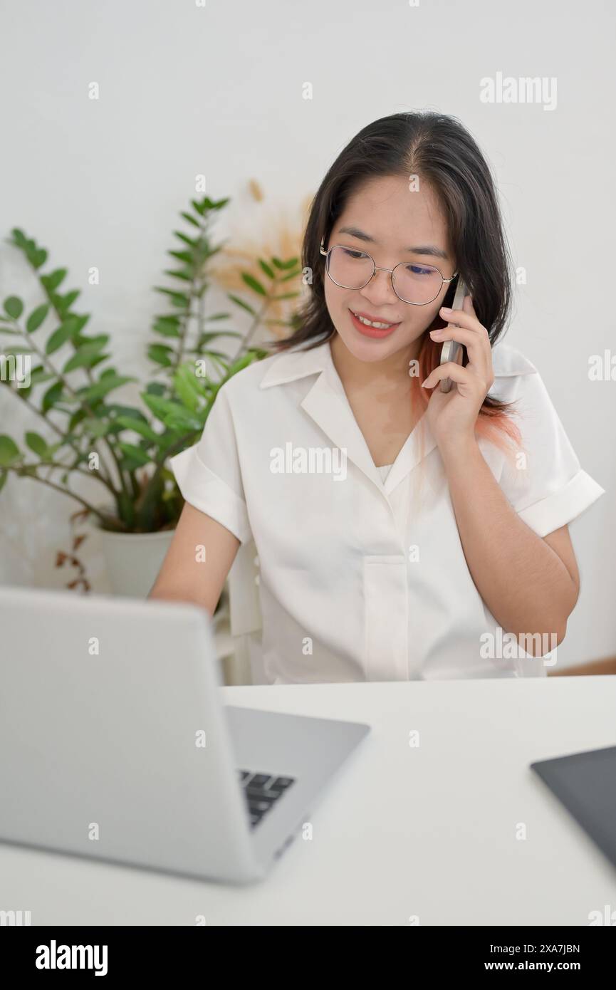 A young positive Asian woman is working remotely from a cafe, talking on the phone with her client while working on her laptop computer. multitasking, Stock Photo