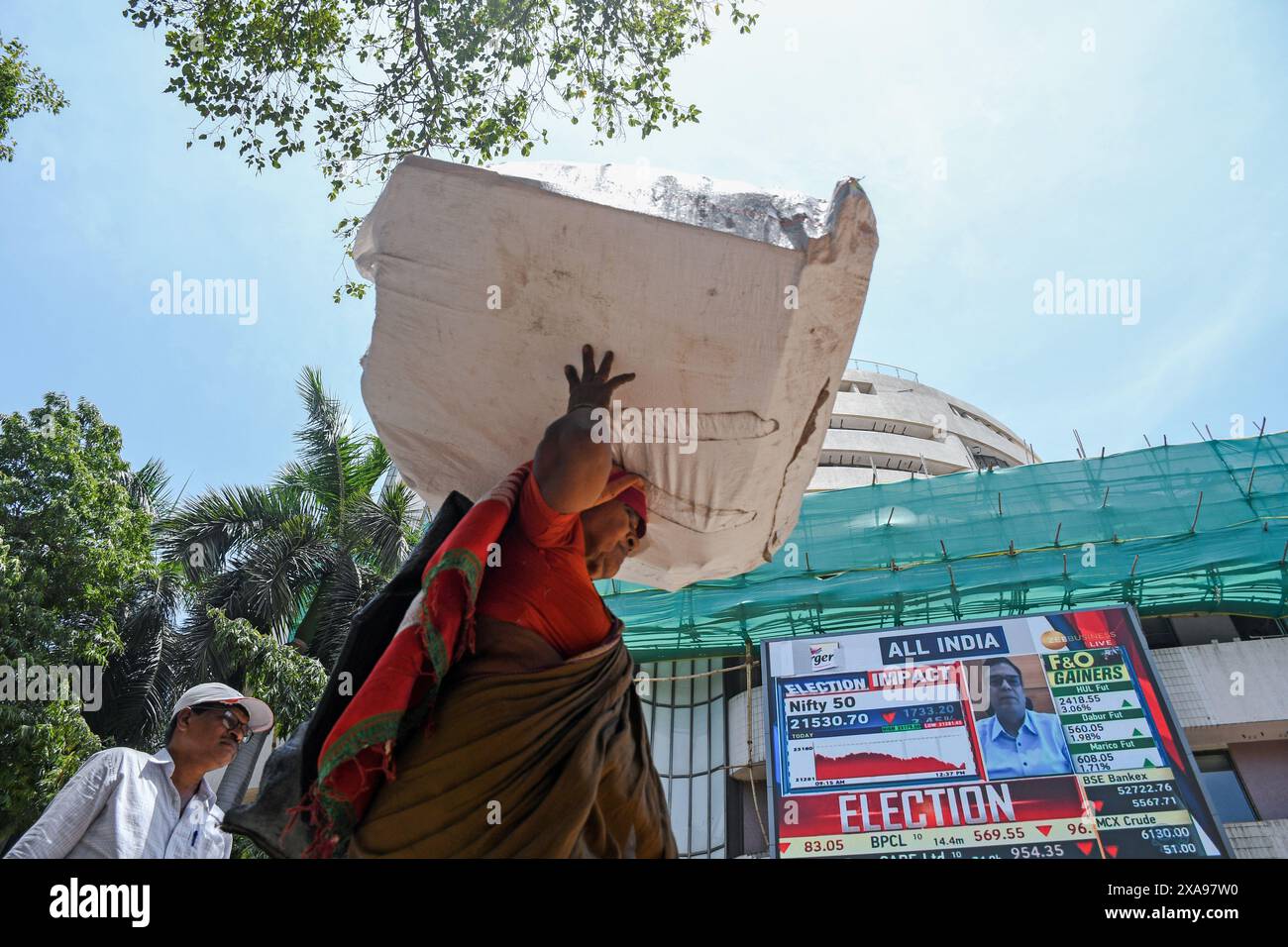 A woman carrying a box walks past the Bombay Stock Exchange (BSE) building as the screen flashes the Loksabha election results and its impact on stock trading. The stock market was volatile when it opened in the morning due to election result, there was an anticipation of Bharatiya Janata Party (BJP) getting the majority of the votes and Prime Minister Narendra Modi getting elected for the third time, but that wasn't the case as opposition parties gave tough fight to the Bharatiya Janata Party (BJP) as votes were being counted and result was flashed on the screen which impacted the stock tradi Stock Photo