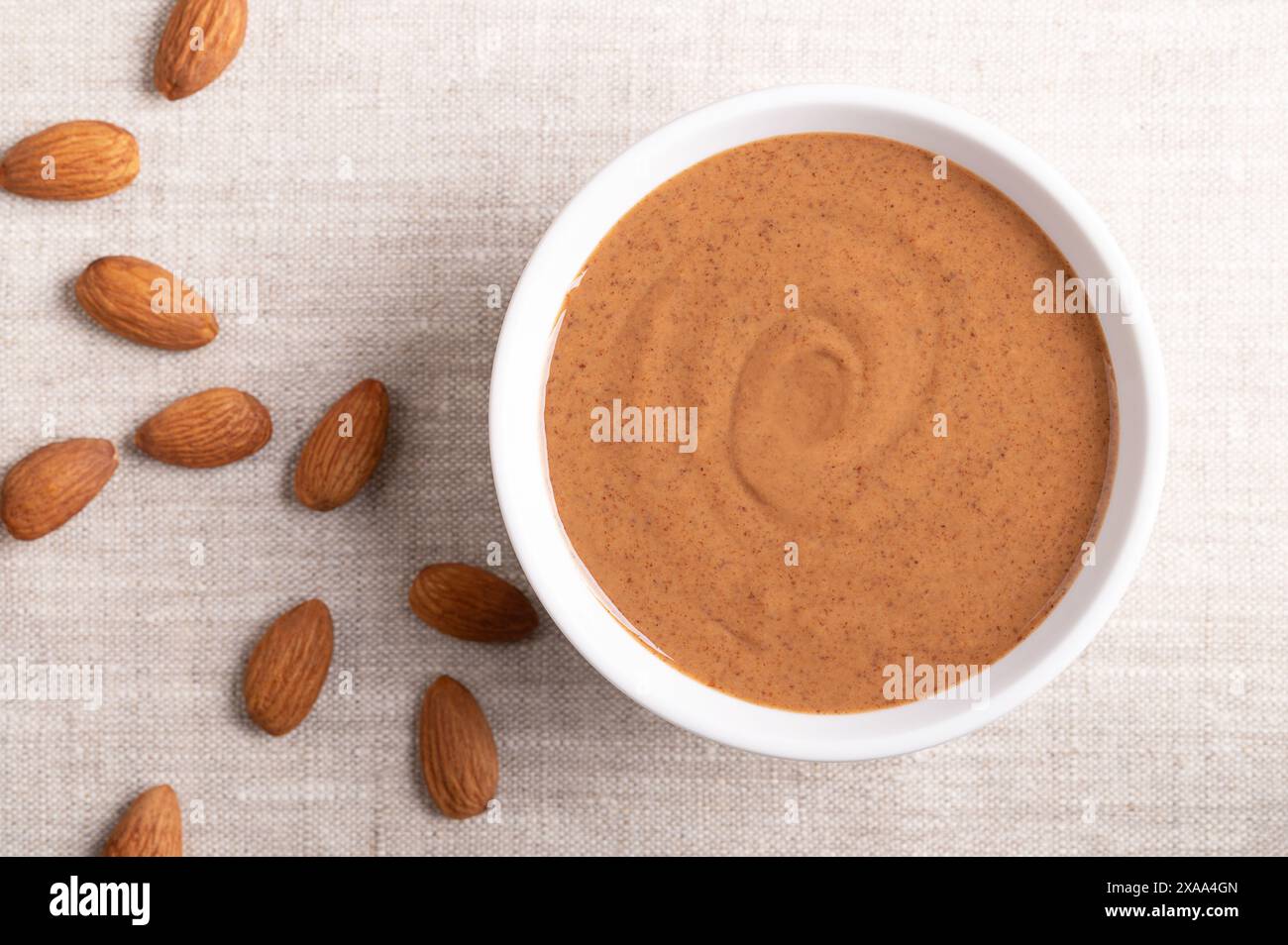 Brown almond butter in a white bowl on linen fabric. Smooth food paste made from grinding almonds into nut butter. Puree of the nuts of Prunus dulcis. Stock Photo