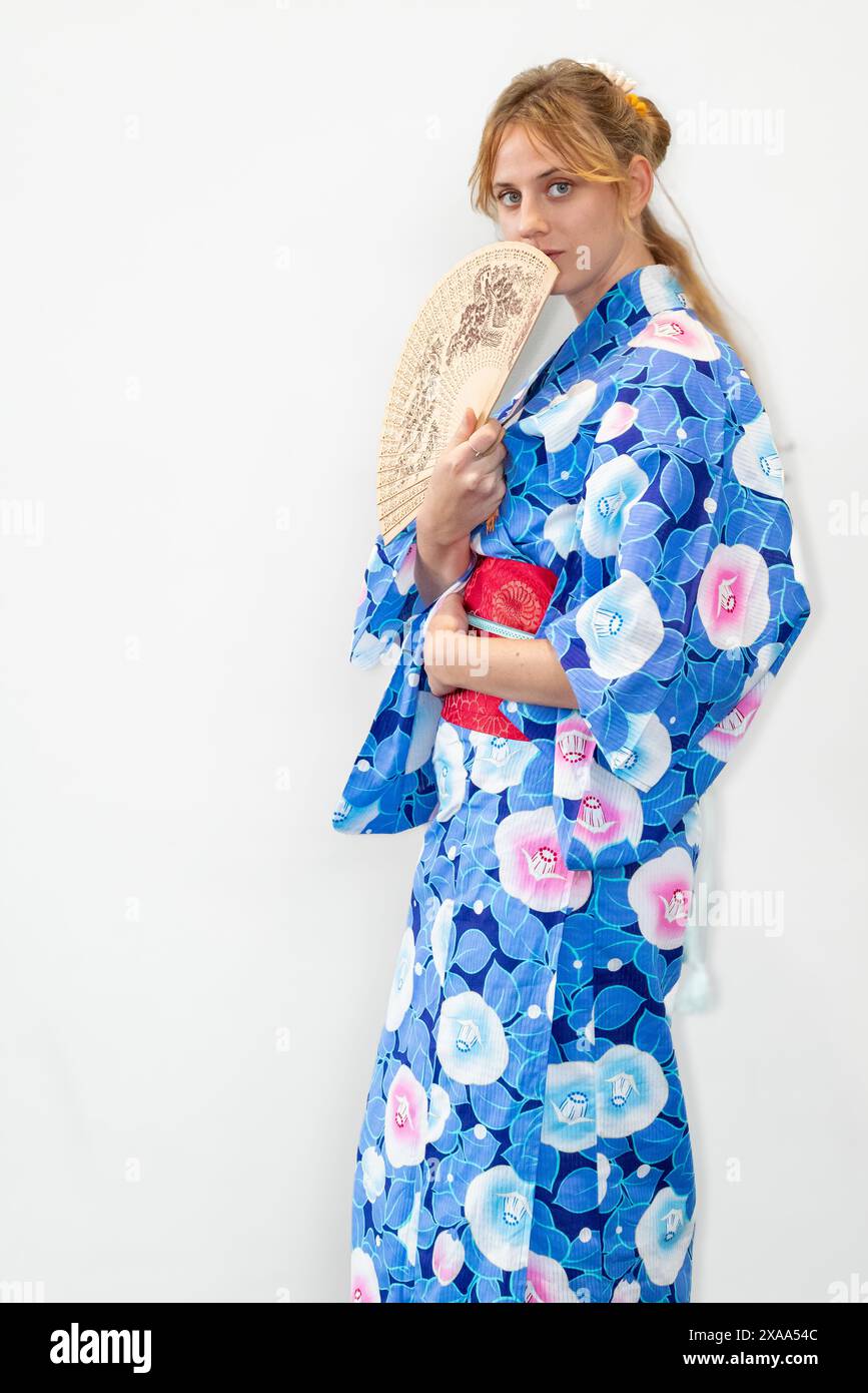 A female cosplayer in a blue kimono with a red sash holding a wooden fan in front of her face Stock Photo