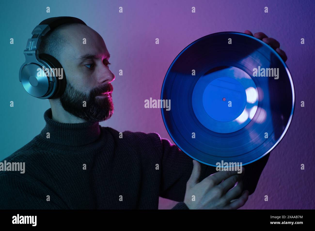 Vintage vinyl disc in hands of Young man with beard in headphones, contemplatively looking in neon purple and green light, Neon-lit contemplation, cri Stock Photo
