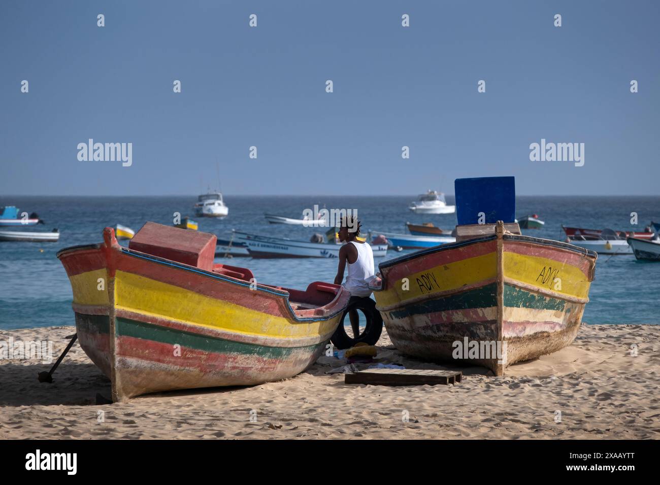 Local Man resting between colourful Fishing Boats on Praia de Santa Maria Beach, Santa Maria, Sal, Cape Verde Islands, Atlantic, Africa Stock Photo