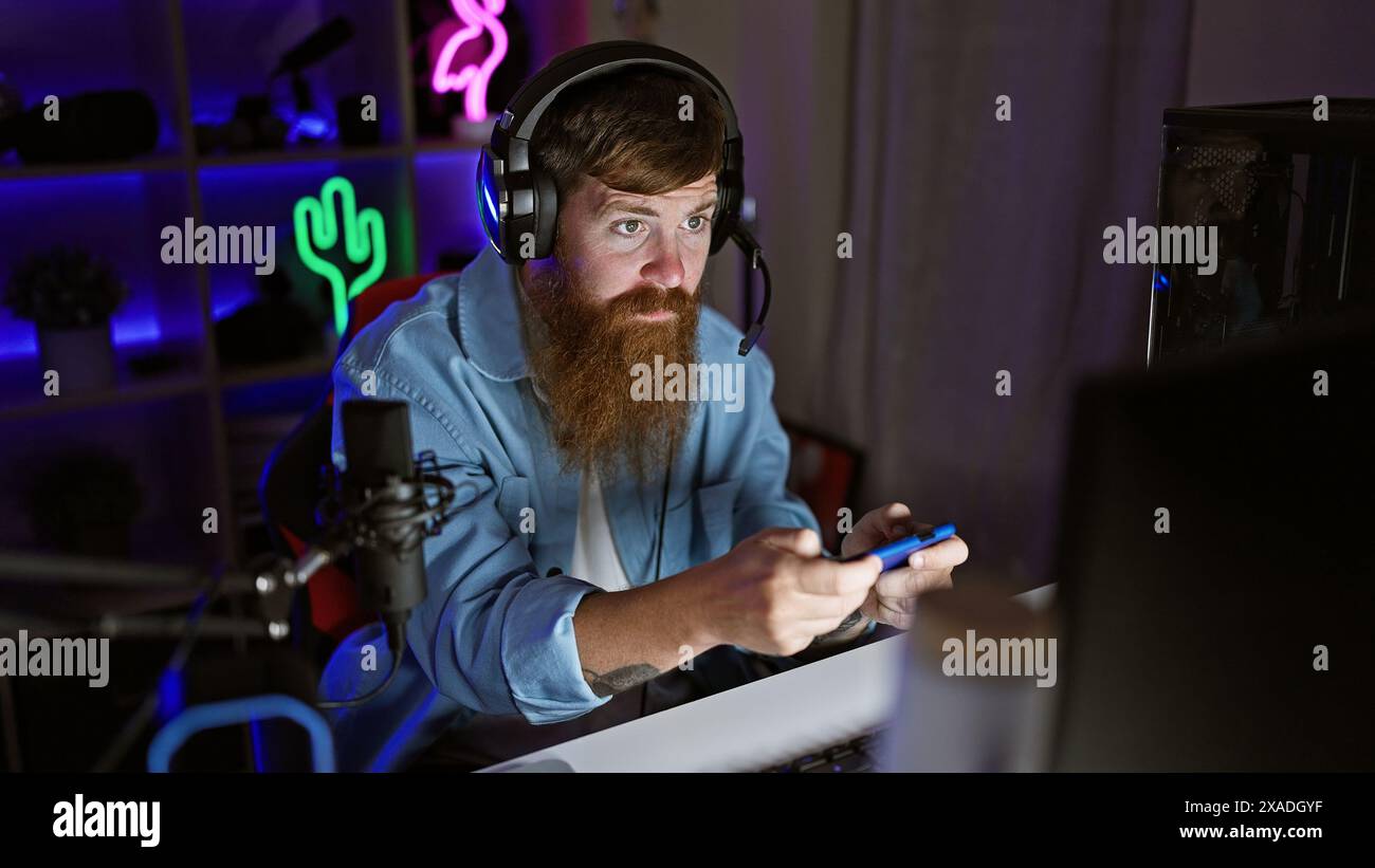Intensely concentrated young redhead man steaming video game on smartphone in dark gaming room at night Stock Photo