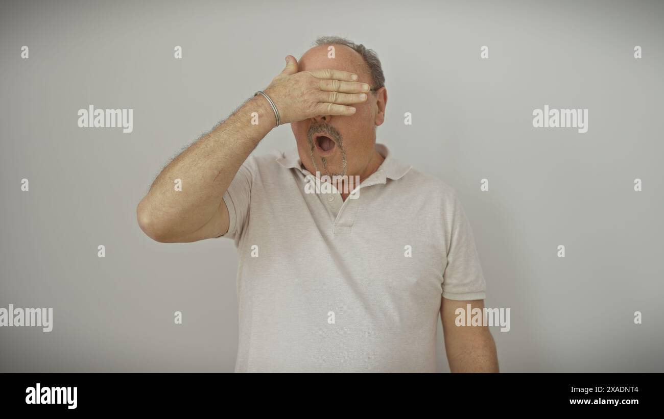 A surprised senior man covers his eyes with his hand against a white ...
