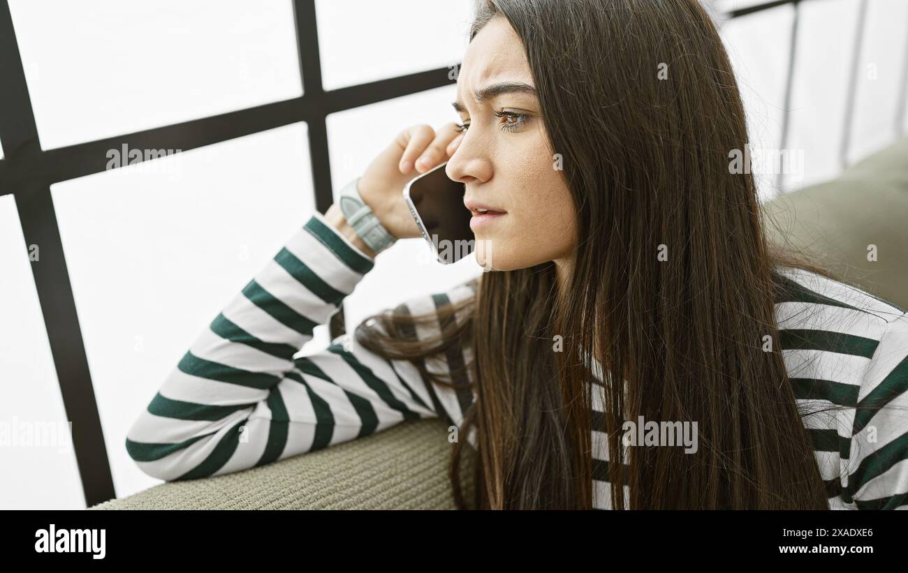 A contemplative young woman on a phone call indoors, exuding a look of concern or deep thought. Stock Photo