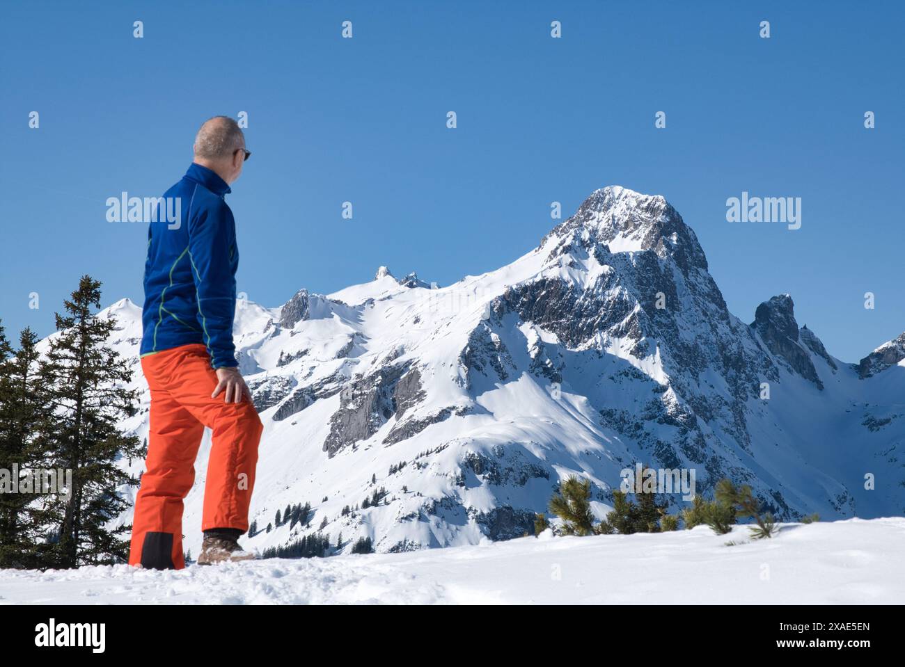 A man in orange pants stands on a snowy mountain, looking out at the beautiful landscape. Concept of adventure and awe, as the man takes in the breath Stock Photo