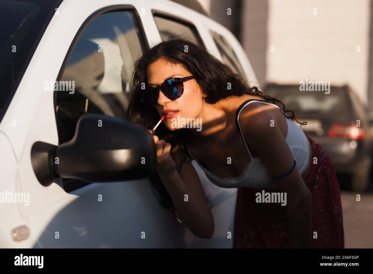 A fashionable young woman in a casual summer outfit uses a car mirror to apply lipstick on a sunny city day, showcasing urban lifestyle and beauty rou Stock Photo