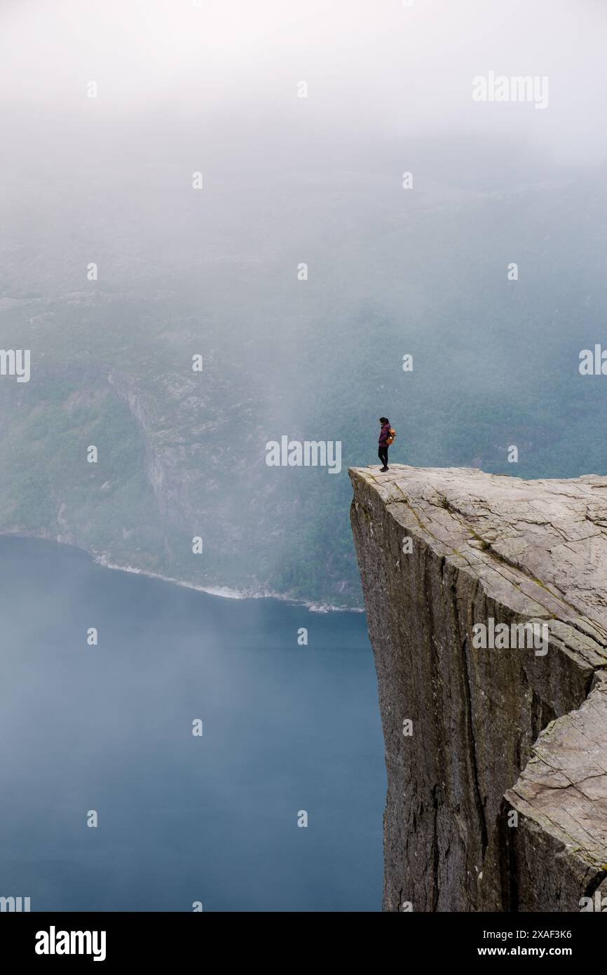 A lone hiker stands at the edge of Preikestolen, a dramatic cliff in Norway. The misty landscape and sheer drop below evoke a sense of awe and solitude. Stock Photo