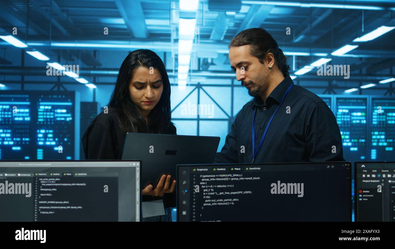 Technicians working in high tech server hub, analyzing data on laptop to ensure seamless data flow. Coworkers examining equipment in data center, ensuring system integrity and security Stock Photo