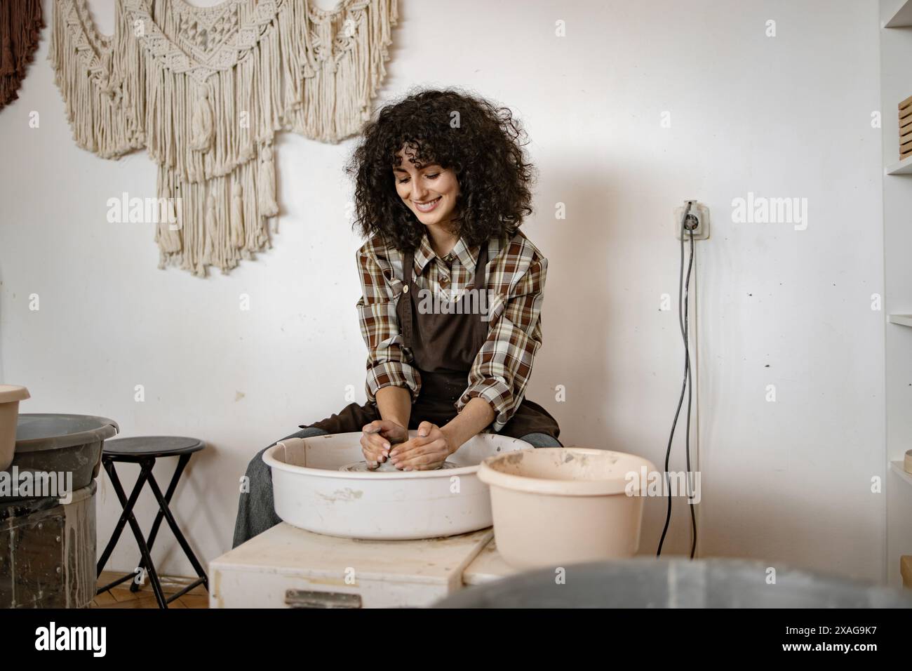 Female potter using wheel to sculpt ceramic bowl in cozy studio. Curly haired woman enjoying creative process. Stock Photo