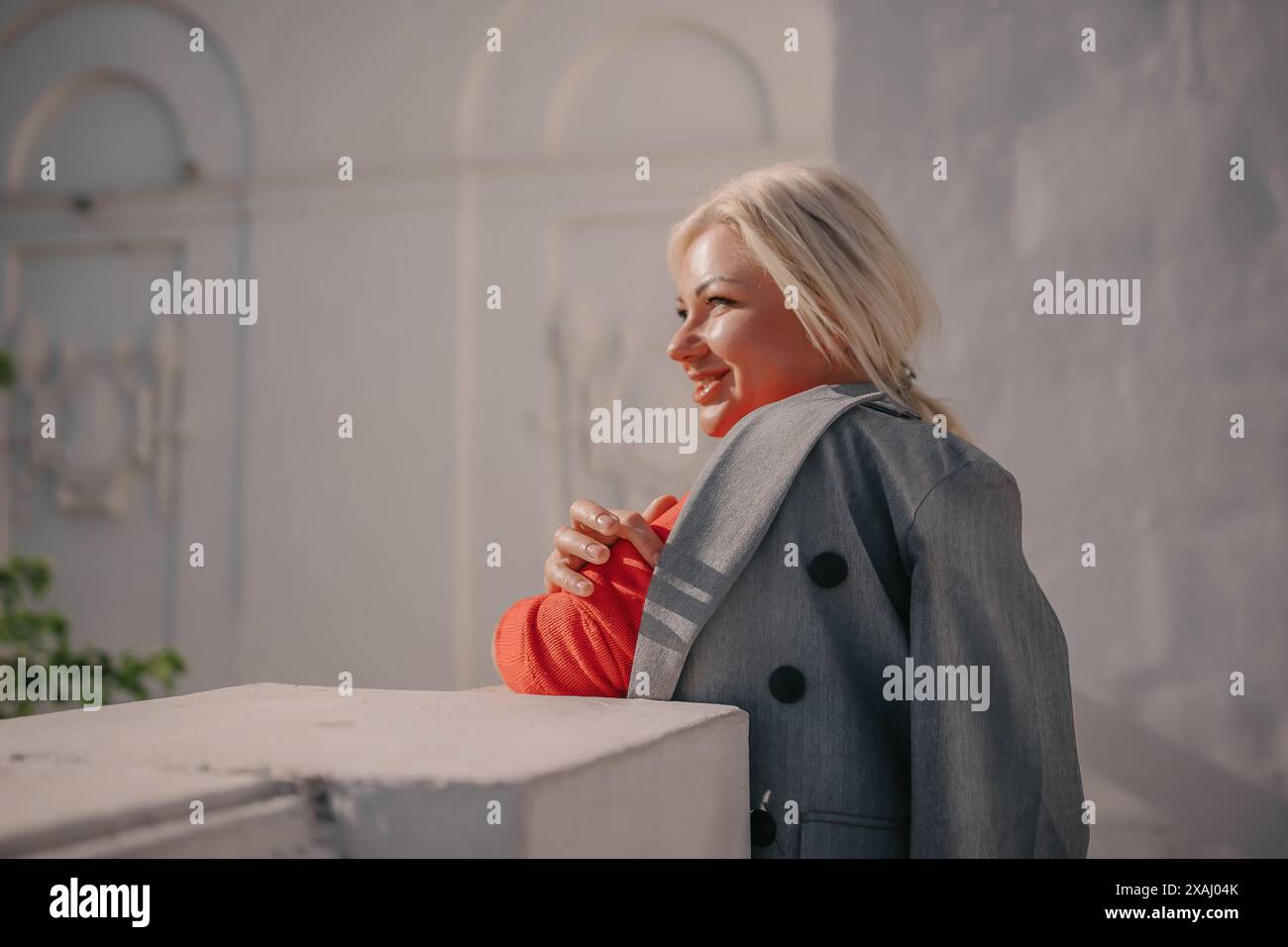 A woman is standing in front of a white building, wearing a gray jacket ...