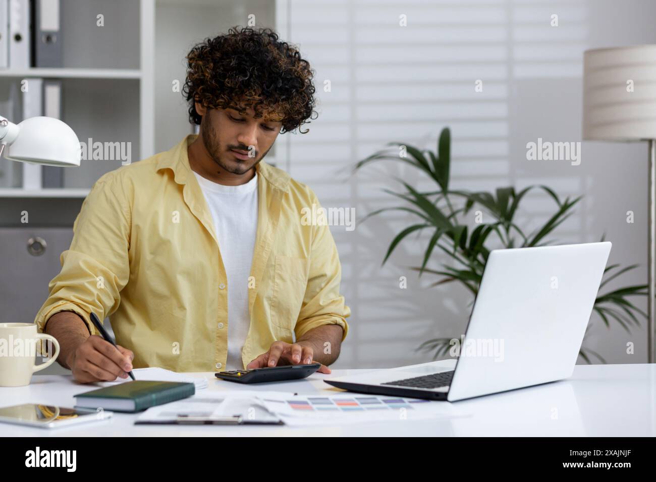 Young man working at home office using a calculator and laptop, analyzing documents and making notes. Stock Photo