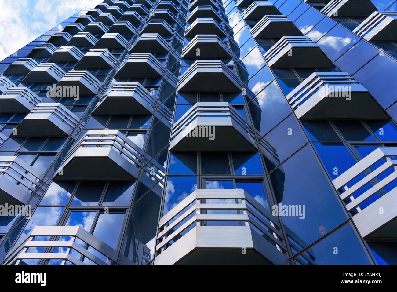 Wide angle upward view of a modern building, showcasing its geometric facade with white balconies and reflective blue glass windows reflecting white c Stock Photo