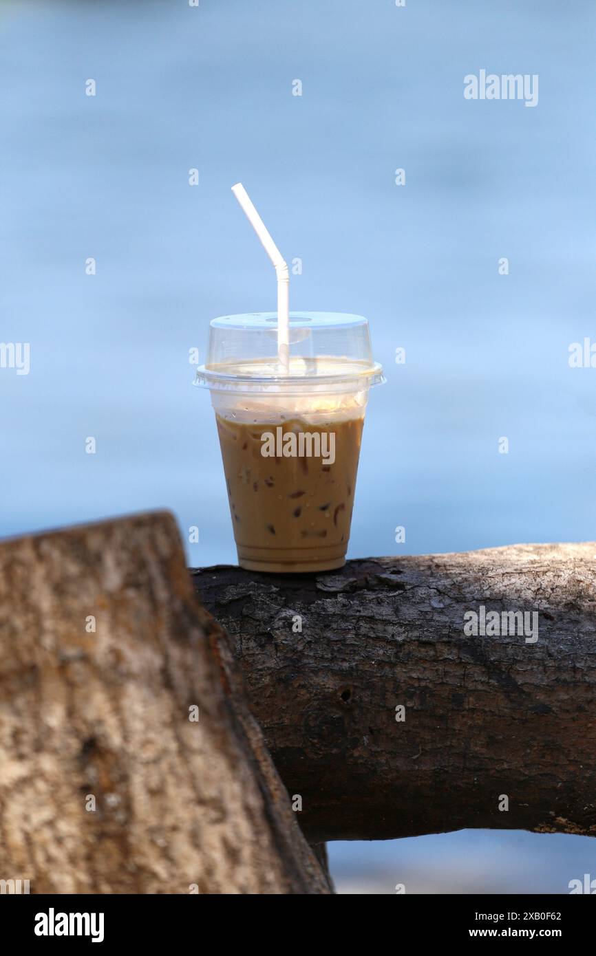 Clear plastic glasses of latte coffee on a big log with the blue sea as ...