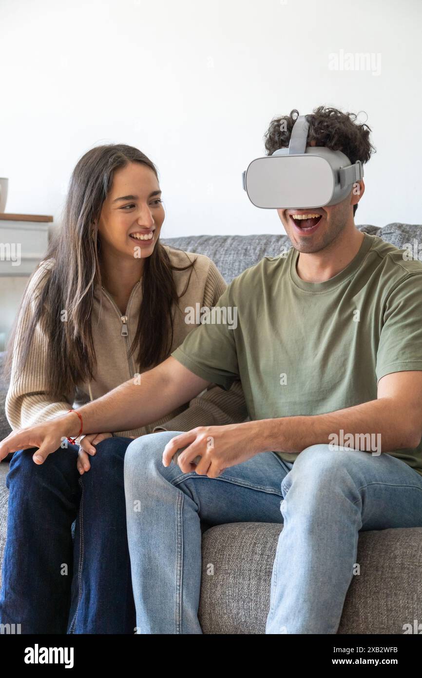 A jovial couple sits on a sofa while the man, wearing a virtual reality ...