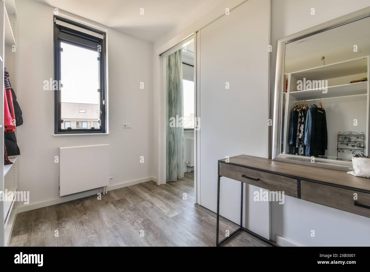 A well-lit modern walk-in closet featuring a built-in wardrobe with open drawers and neatly arranged clothes, complemented by a smooth wooden desk and Stock Photo