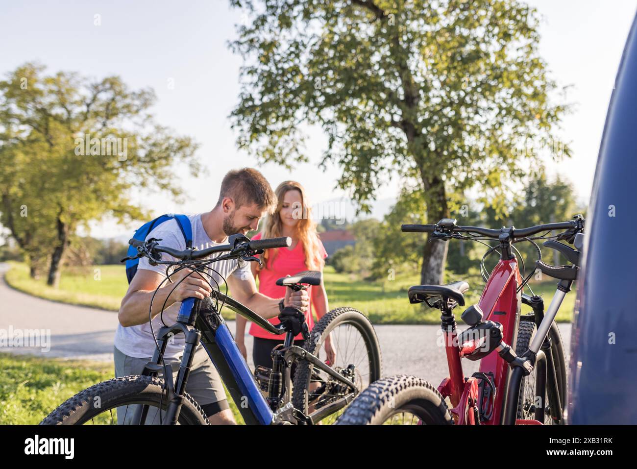 Young man and a woman preparing for off-road biking, taking down electric mountain bikes from the bike rack on the camper van. Stock Photo