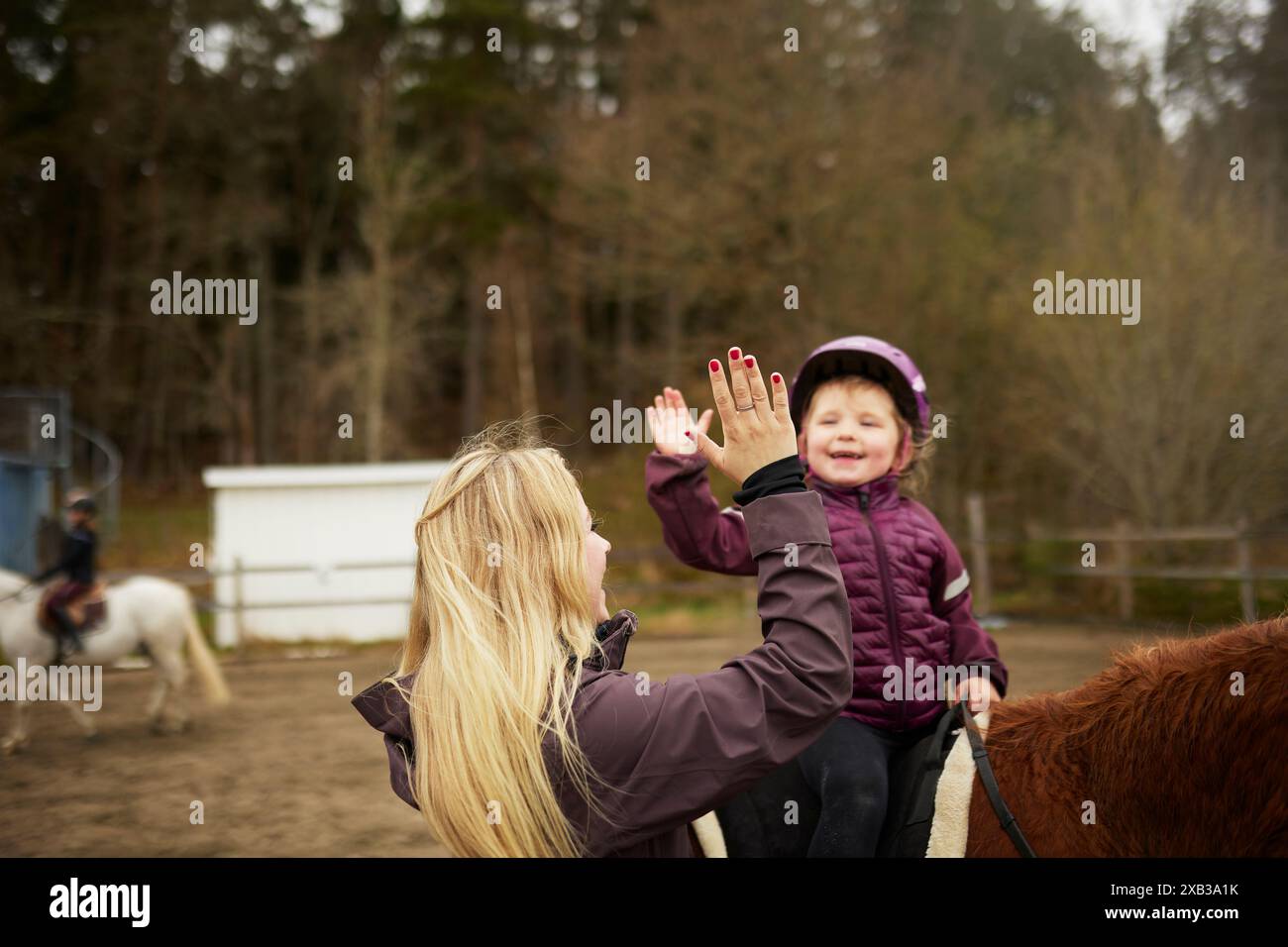 Rear view of blond female instructor giving high-five to boy sitting on horse at ranch Stock Photo