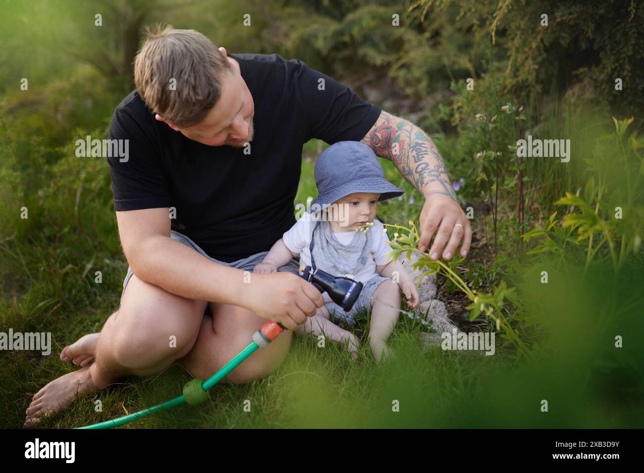 Father spraying water while sitting with toddler son in back yard garden Stock Photo