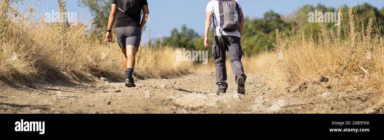 Cinematic cropped photo of two hikers walking on a dirt road in the mountains, back view of two guys walking uphill on the mountain roads, hikers walk Stock Photo