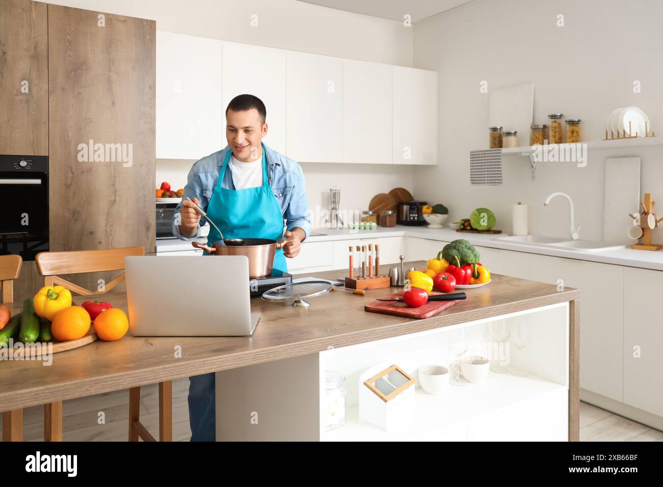 Young man with laptop cooking soup in kitchen Stock Photo