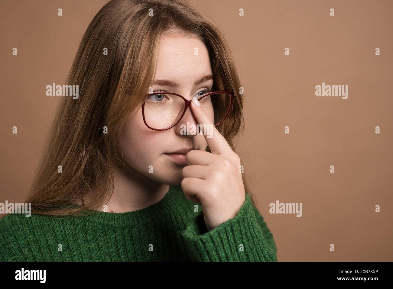 A young woman in a green sweater and glasses is gently touching her nose, showcasing elements like vision care, human body, and eyewear Stock Photo