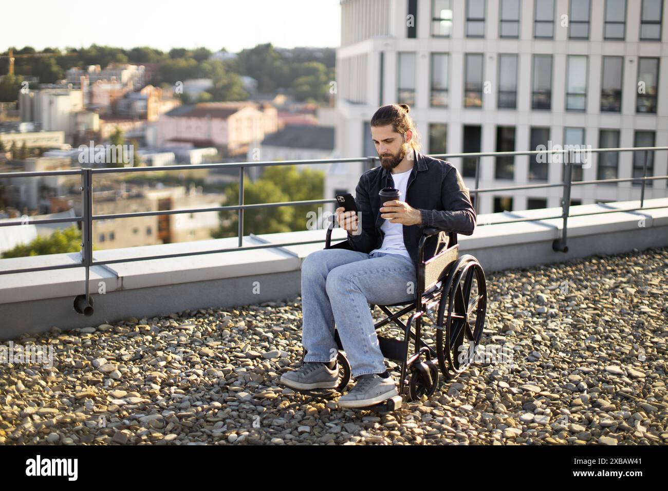 Man in wheelchair using phone and drinking coffee on rooftop Stock Photo