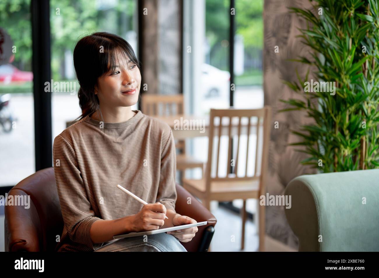 An attractive Asian woman sits in an armchair with her digital tablet ...