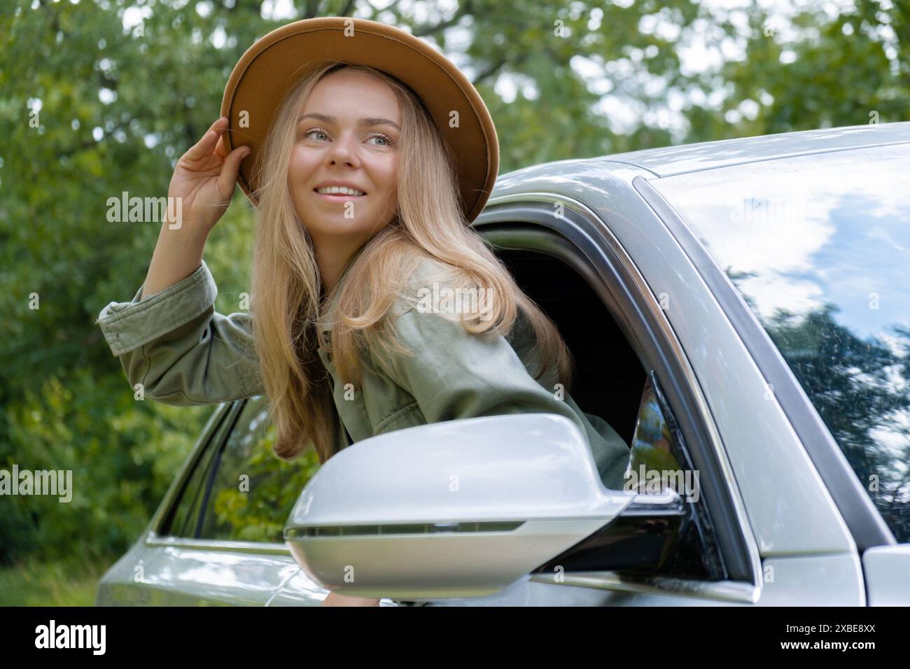 Blonde woman in hat sticking head out of windshield car. Young tourist explore local travel making candid real moments. True emotions expressions of getting away and refresh relax on open clean air Stock Photo