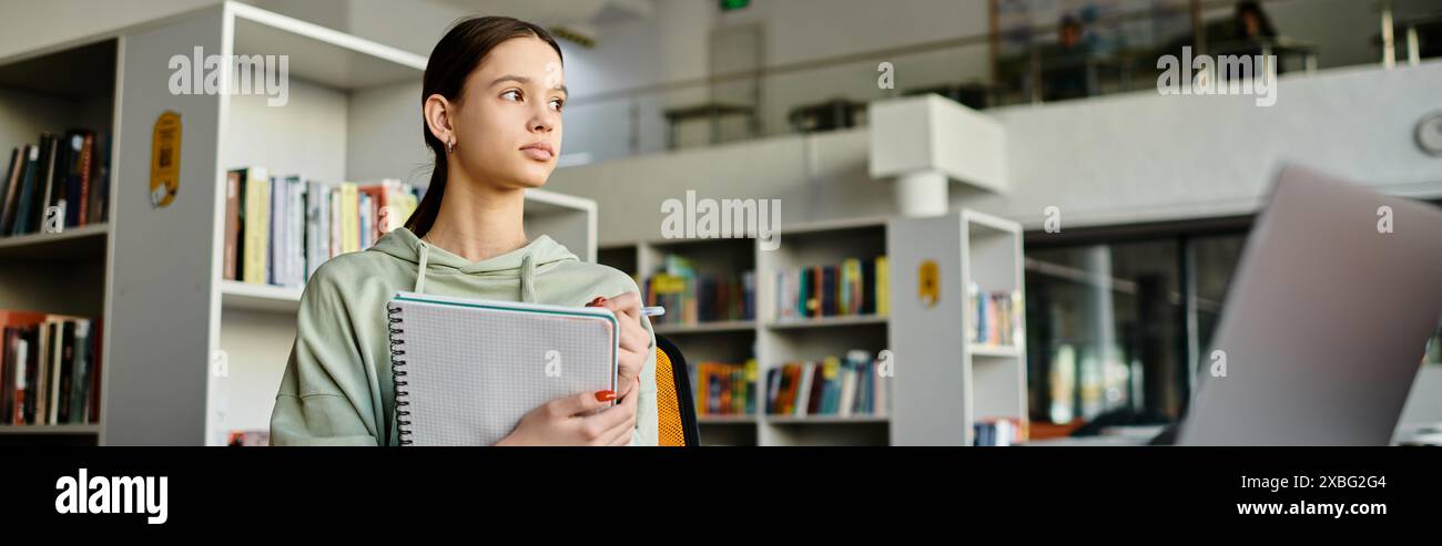 A teenage girl stands in front of a bookcase in a library, immersed in studying and doing homework on her laptop after school. Stock Photo