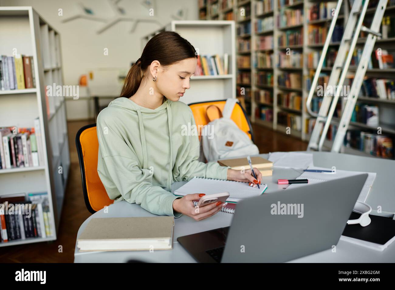 A teenage girl is focused on her laptop at a table in a library, engaging in modern education after school. Stock Photo