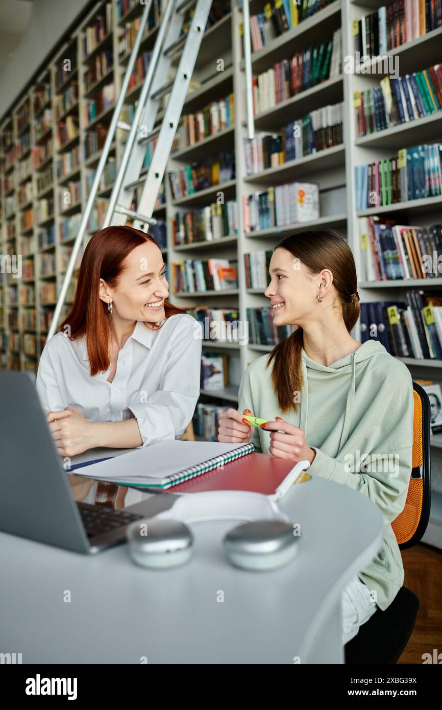 A redhead tutor is teaching a teenage girl in a library, engaging in after-school lessons while using a laptop. Stock Photo