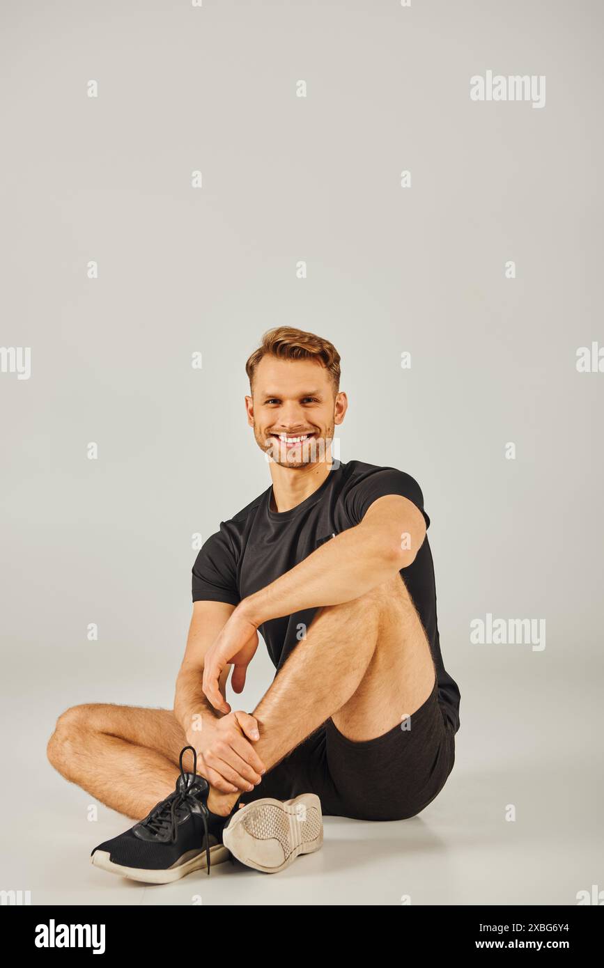 A young athletic man in active wear sitting on the floor and smiling cheerfully in a studio with a grey background. Stock Photo