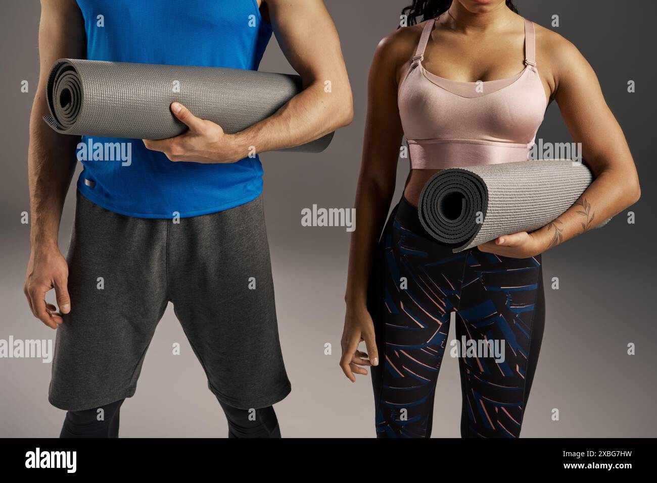 A young multicultural and fit couple in active wear holding a yoga mat in a studio setting against a grey background. Stock Photo