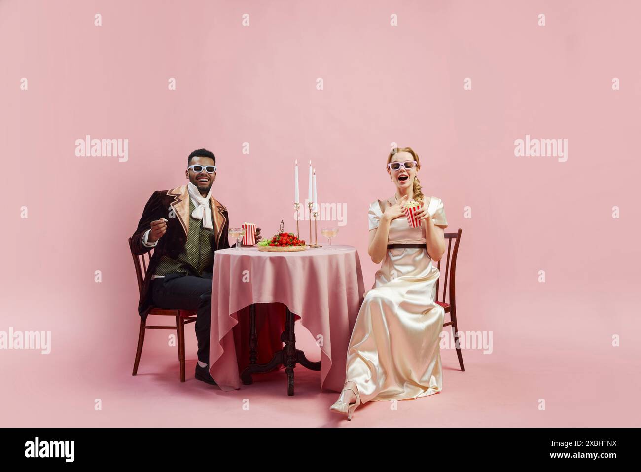 Elegant Victorian couple in period costumes sitting at table and watching movie in 3D glasses, with popcorn against pink studio background Stock Photo