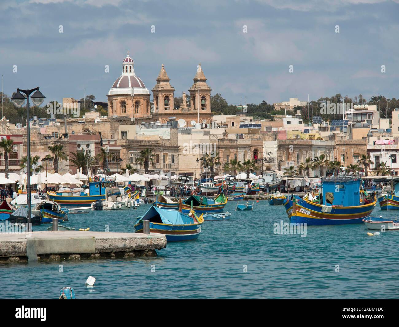 Boats in the harbour with an urban backdrop and a cathedral under a cloudy sky, marsaxlokk, mediterranean sea, malta Stock Photo