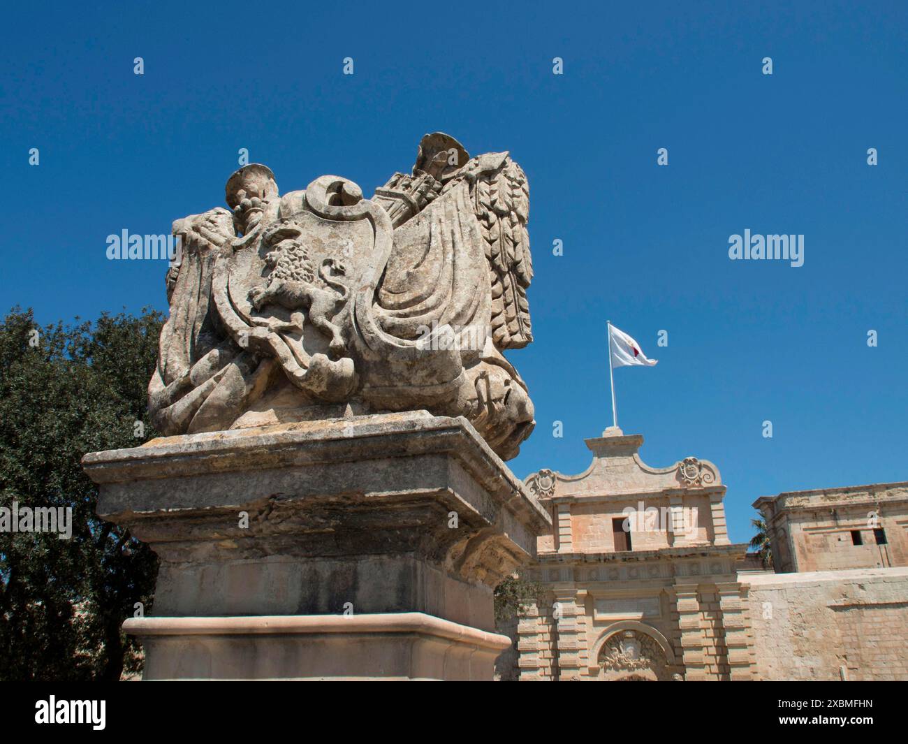 Close-up of an ornately carved historical statue with a coat of arms and a flag in the background, mdina, mediterranean sea, Malta Stock Photo