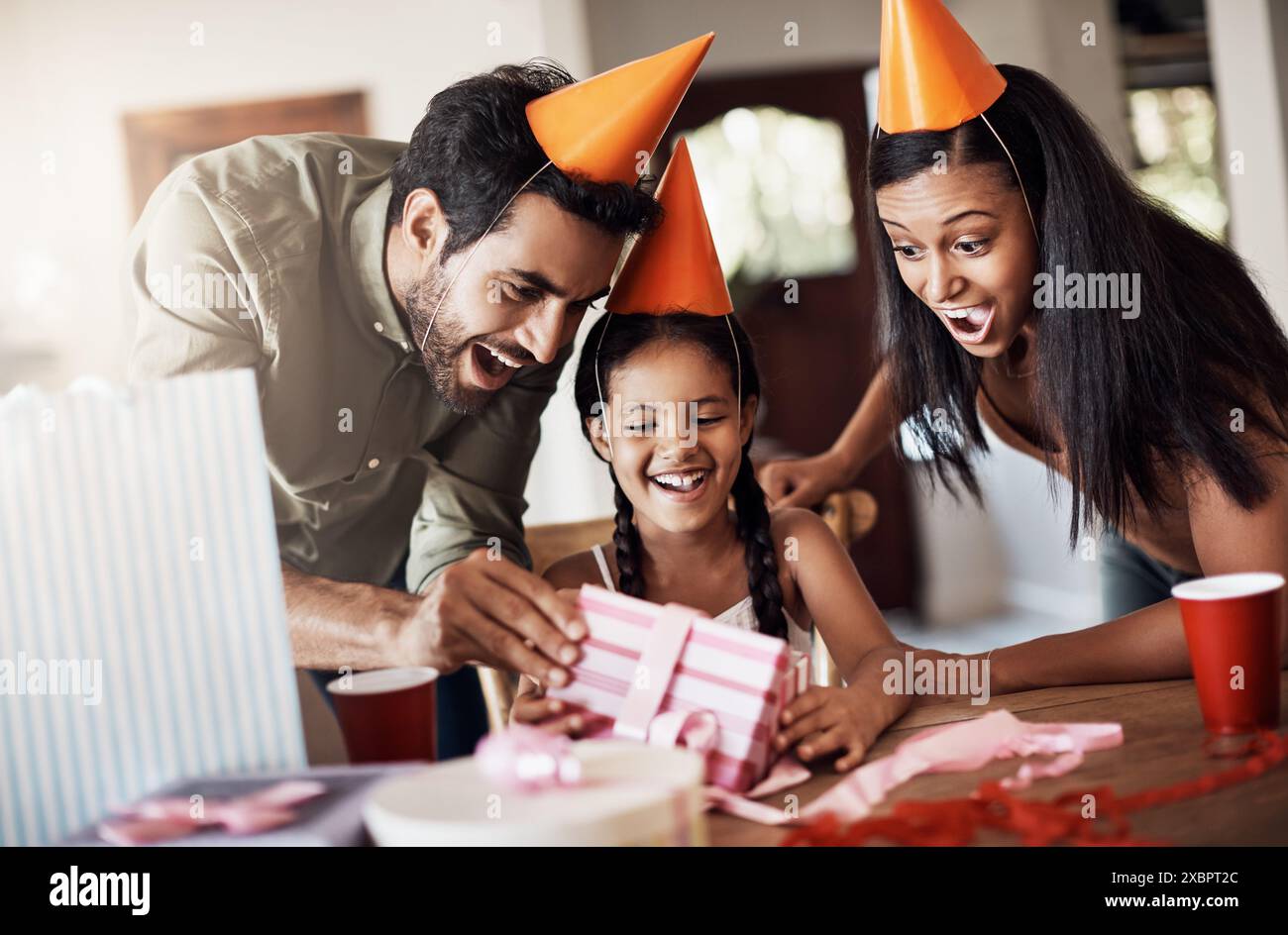 Parents, child and wow in home for birthday gift in table with party hat and fun. People, family and happy or excited for present with love, care and Stock Photo