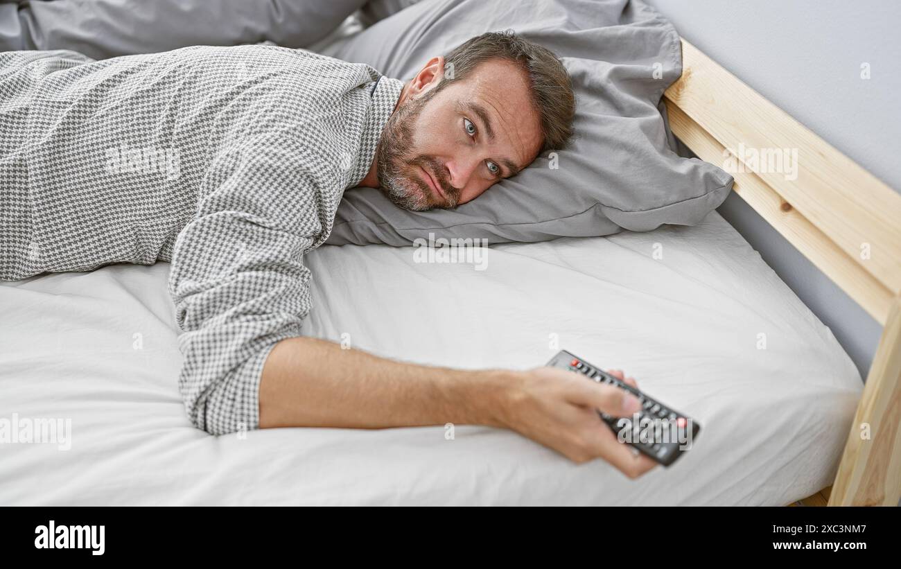 Middle-aged hispanic man with beard lying in bed holding remote control indoors. Stock Photo