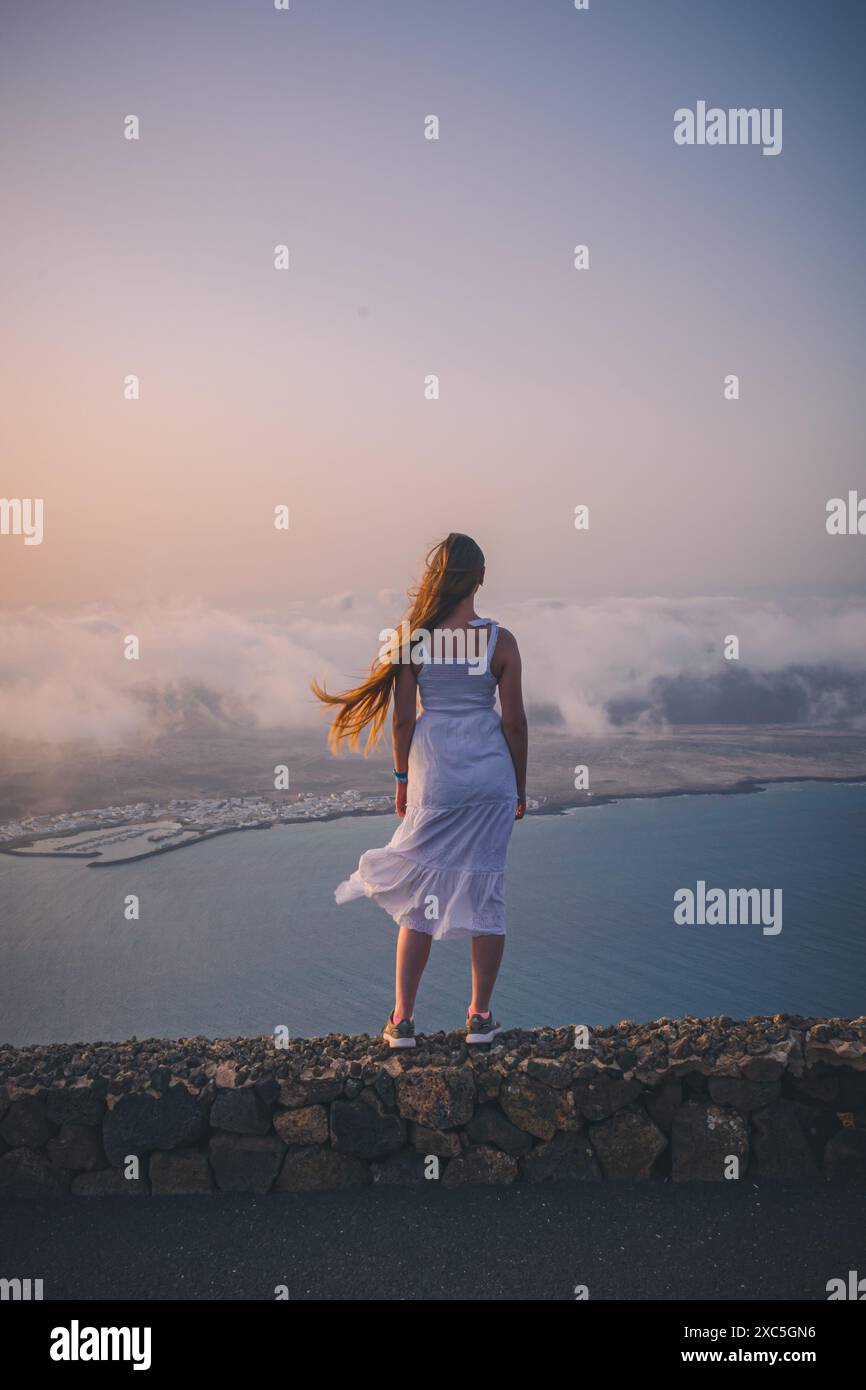 Young woman in white dress stands on a stone wall and looks on a island ...