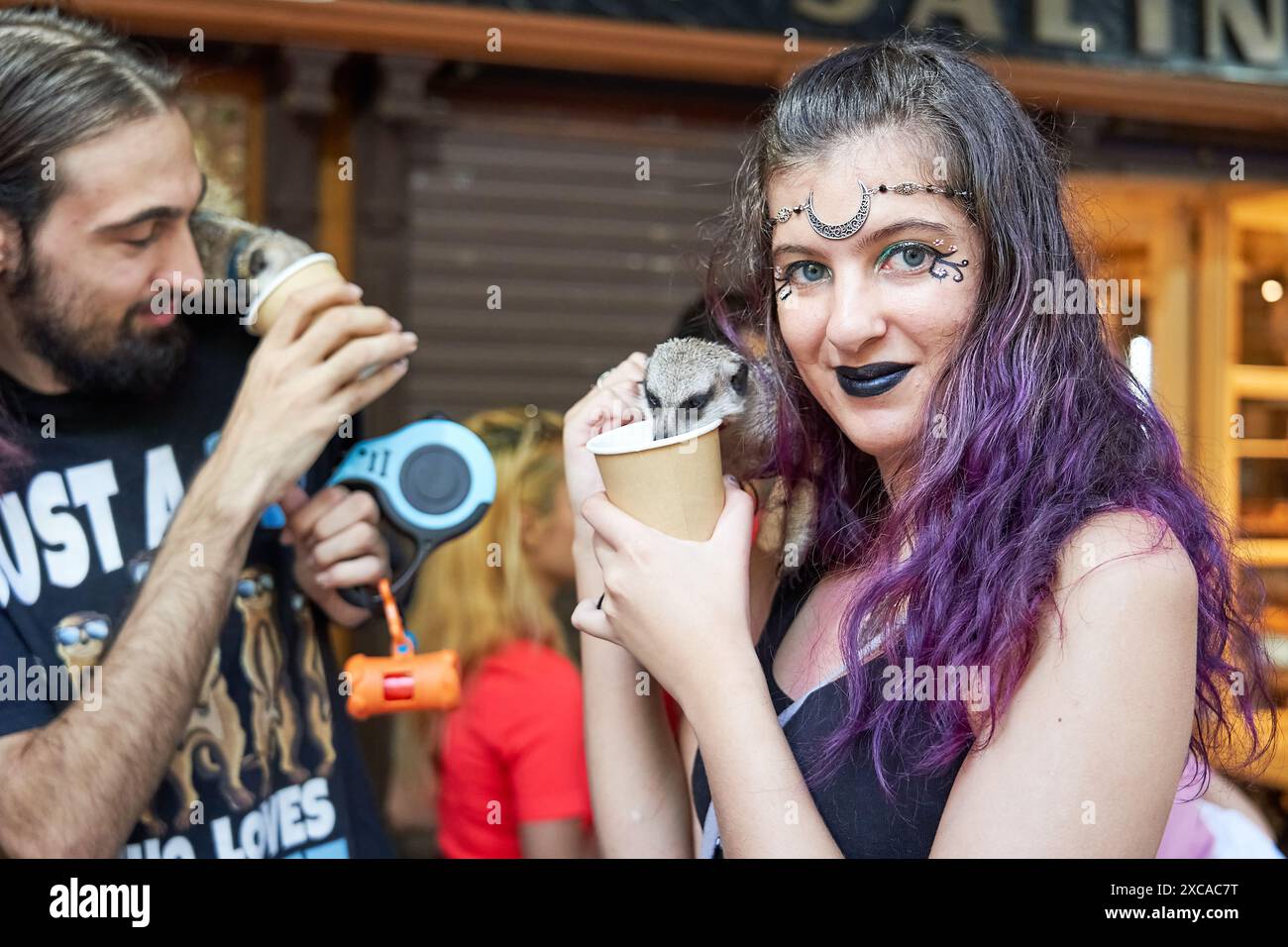 A couple with their pet ferrets on their shoulders letting them drink from cups. She has purple violet hair and a metal headband and green lipstick Stock Photo