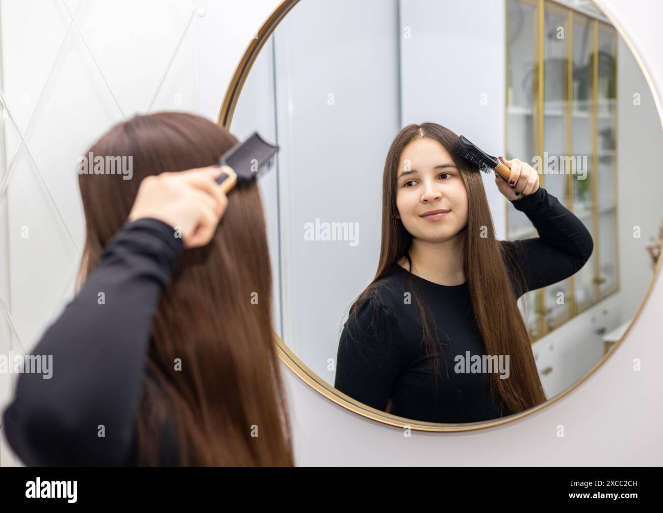 Young woman in a black long sleeve brushes her long hair with a hairbrush while looking in the mirror. Stock Photo