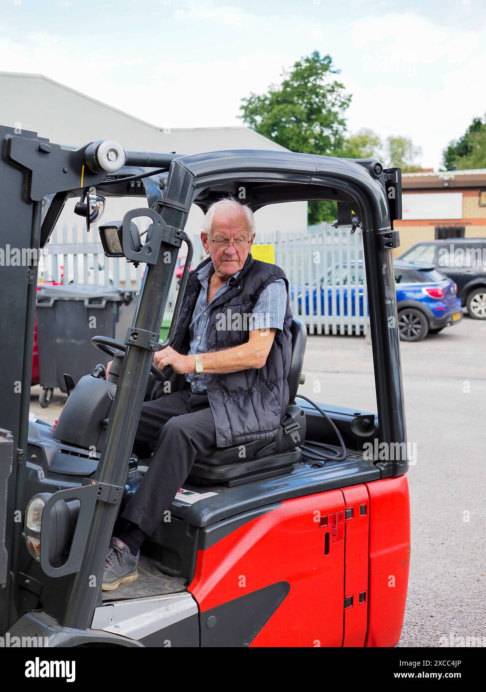 Elderly man 80-90 operating a red forklift in an industrial yard on a sunny day, focused and determined. Stock Photo